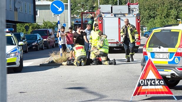 Brandmän och polis på olycksplats, arbetar med någon på marken. En trafikolycka har inträffat och flera räddningsfordon står parkerade runt omkring.