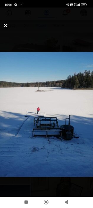 Snötäckt brygga med en person i röd jacka som går på frusen sjö omgiven av skog under klarblå himmel.