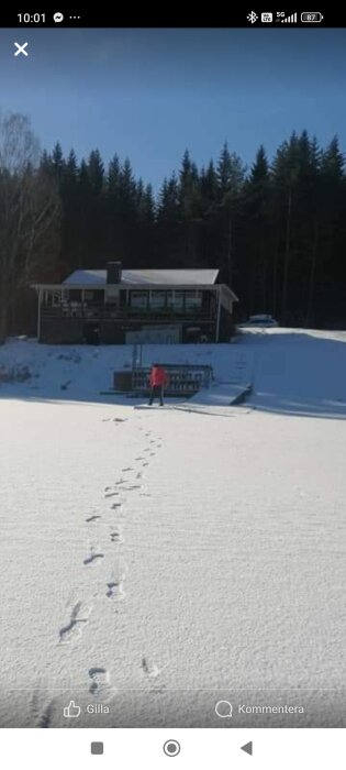 Ett snötäckt landskap med ett hus i bakgrunden och fotspår som leder till en person i röd jacka.