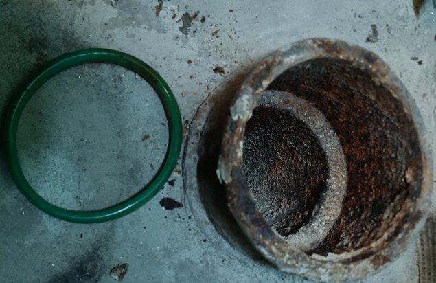 Gammalt avloppsrör av gjutjärn med rost inuti och en grön gummiring till vänster liggande på betonggolv, i behov av tätning.