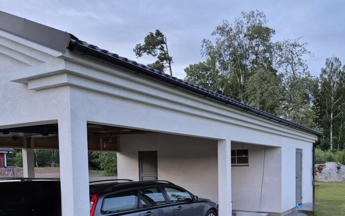 Vit putsad carport med svart tak och synliga stuprännor, där en grå bil är parkerad, omgiven av träd med en molnig himmel i bakgrunden.