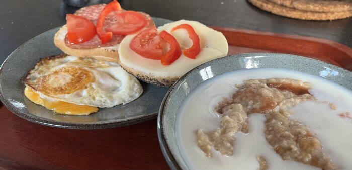 Frukost med havregrynsgröt med sylt och mjölk i en skål, en tallrik med stekt ägg och vallmobräck med ost, tomat och salami.