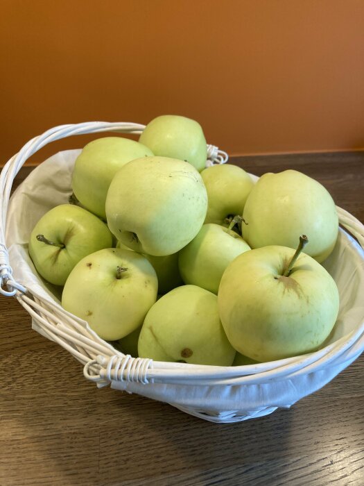 Korg med gröna äpplen, möjligtvis transparente blanche, på ett bord med orange vägg i bakgrunden.