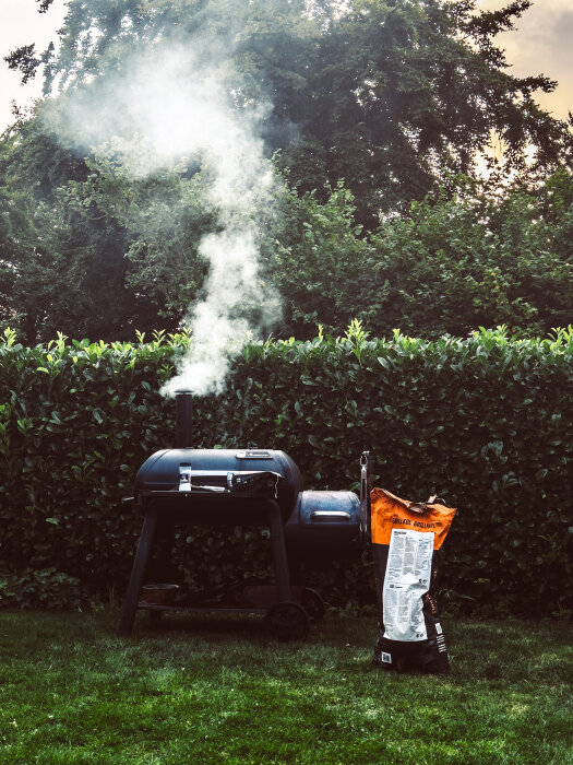 Rök kommer från en svart grill med ett paket ICA grillkol bredvid, och bilden är tagen framför en grön häck.