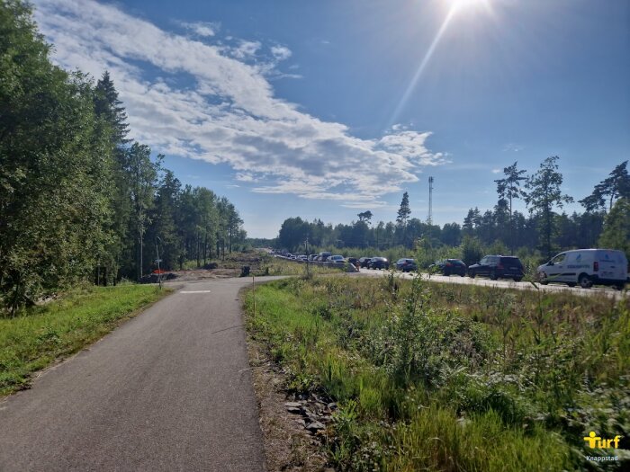 En asfalterad gångväg i natur med blå himmel och sol, skog på vänster sida och bilar som köar på höger sida där det planeras en cykelbana.