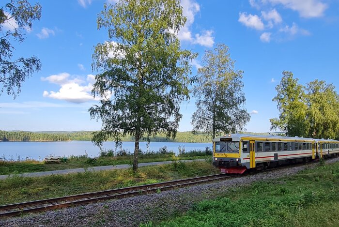 Rälsbuss i Dalsland kör längs en sjö med grönskande träd i bakgrunden under en klarblå himmel.