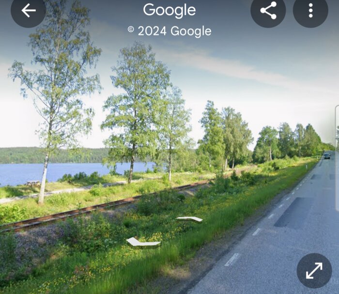 En landsväg vid en sjö kantad med träd med järnvägsspår parallellt med vägen, sommartid under klar himmel.