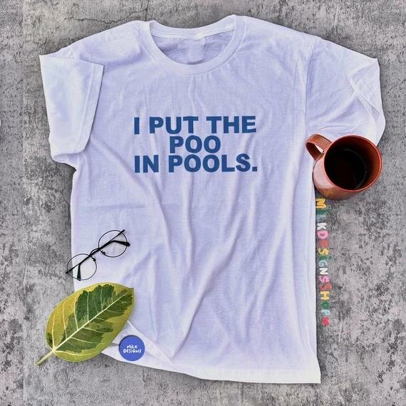 Vit T-shirt med texten "I PUT THE POO IN POOLS." Ligger på betong med en kaffemugg, ett par glasögon och ett blad bredvid.