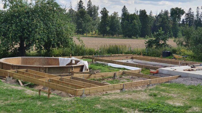 Pågående byggnation av altan runt en pool med trästommar och hål för murstenar i en trädgård med natur i bakgrunden.