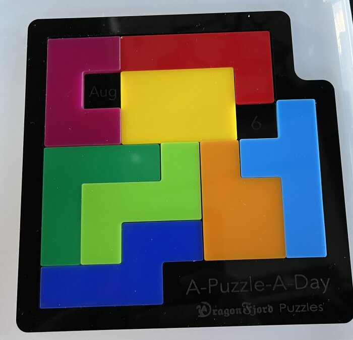 Flera färgglada pusselbitar i olika former är placerade i en pusselram med texten "A-Puzzle-A-Day" och "DragonFjord Puzzles".