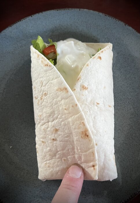 En tortilla-wrap fylld med kycklinggryta, grönsaker och vitlökssås ligger på en grå tallrik. Ett finger håller i botten av wrapen.