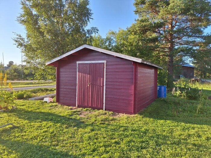 Rödmålad bod med dörr och vindskivor i kontrasterande färg, omgiven av grönska på en solig dag.