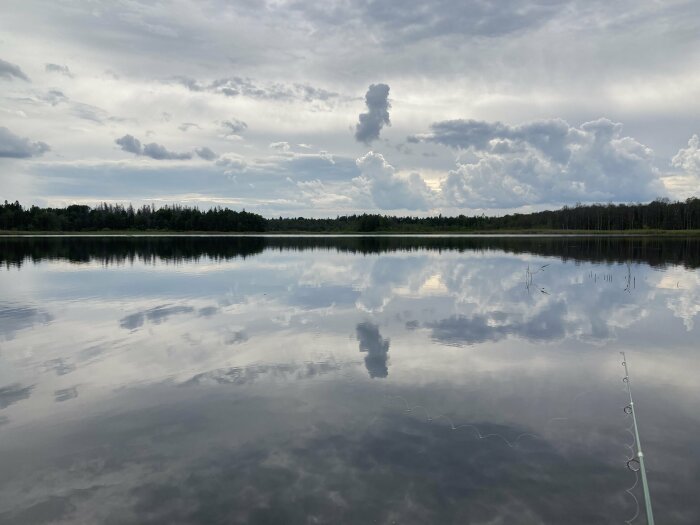 En stilla sjö med spegelblankt vatten reflekterar himlens moln och skogen vid horisonten, med ett fiske- eller metspö som sträcker ut från högerkanten.