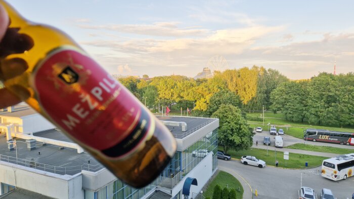 En person håller en Mez'pils-ölflaska med utsikt över en grön park och en stadssilhuett i Riga i bakgrunden.