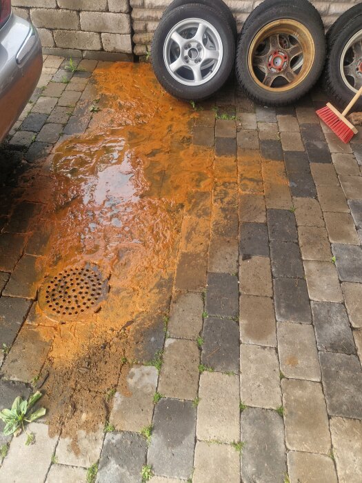 En uppfart med kantarellgul sediment och vattenläckage nära grund av ett hus, tre däck och en röd borste i bakgrunden.