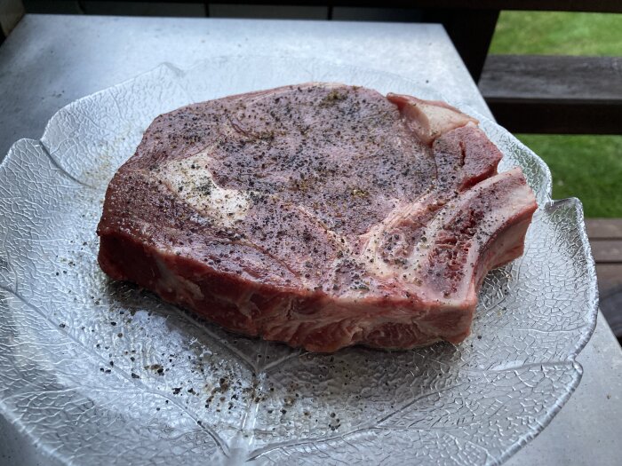 En rå, kryddad Cote de boeuf-biff ligger på ett glastallrik utomhus, redo för grillning.