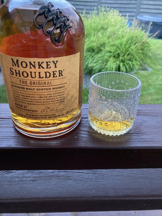 Flaska med Monkey Shoulder whisky och ett halvt fyllt glas står på en träbänk. Grönska och ett staket syns i bakgrunden.