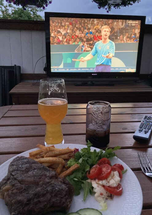 Middag med grillat kött, pommes, sallad, ett glas öl och en dricka framför en TV som visar en bordtennismatch.