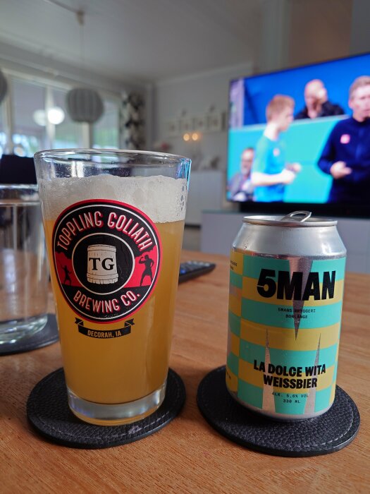 En burk med 5Mans La Dolce Wita Weißbier och ett glas med öl på ett bord, med en TV som visar en sporthändelse i bakgrunden.