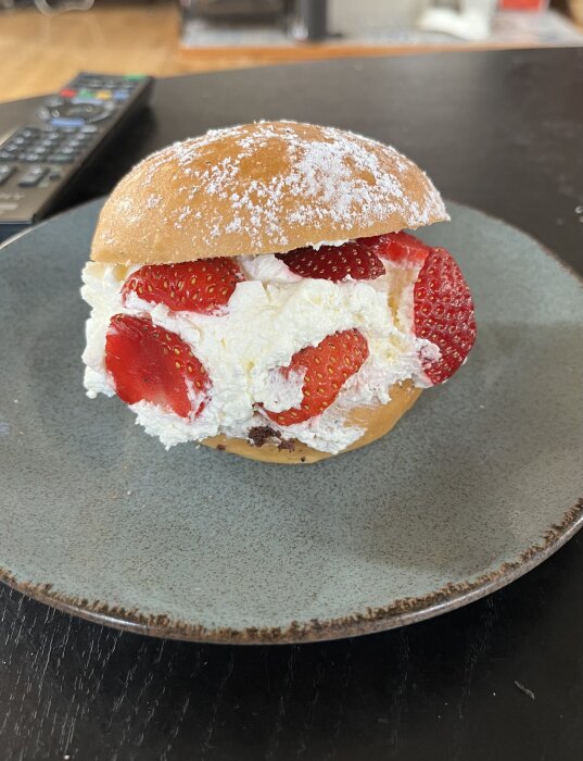 En bakelse med florsocker på toppen, fylld med grädde och jordgubbar, på en grå tallrik. En fjärrkontroll syns i bakgrunden.