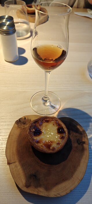 Ett glas dessertvin och en portugisisk pasteis de nata på ett runt träfat, stående på ett ljust träbord.