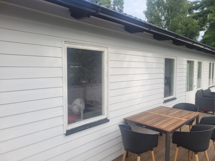 Vitmålad fjällpanel på ett hus med svarta takrännor och ett träbord med fyra stolar på verandan.