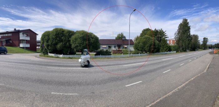 En genomfartsväg med trottoar bredvid ett bostadsområde. Ett hus med träd och en vit staket syns, och en motorcykel med sluten förarhytt passerar.