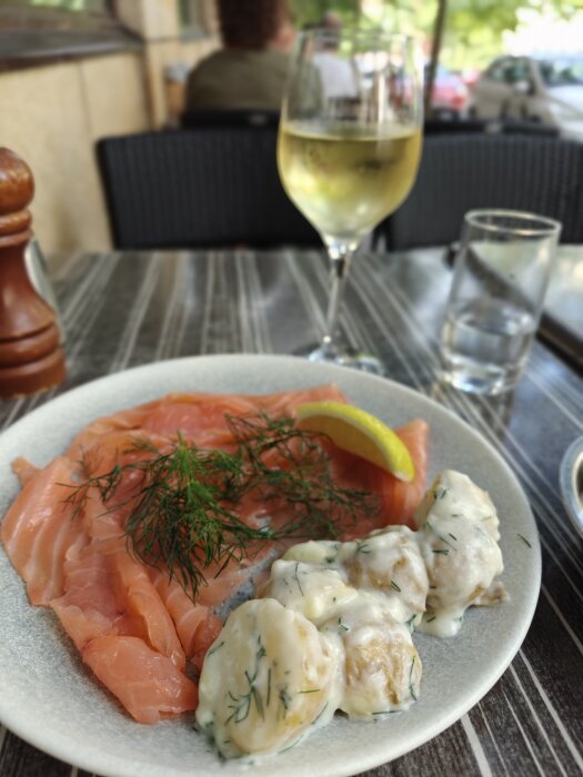 Tallrik med gravad lax och dillpotatis samt ett glas vitt vin på ett härligt sommarcafé.