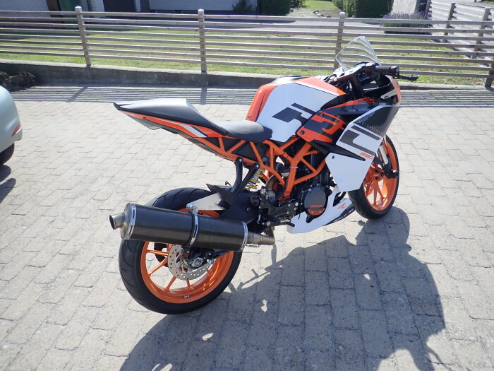 Motorcykel med orange och svart färgschema, av modellen KTM 390, parkerad på en stenlagd yta bredvid ett staket.