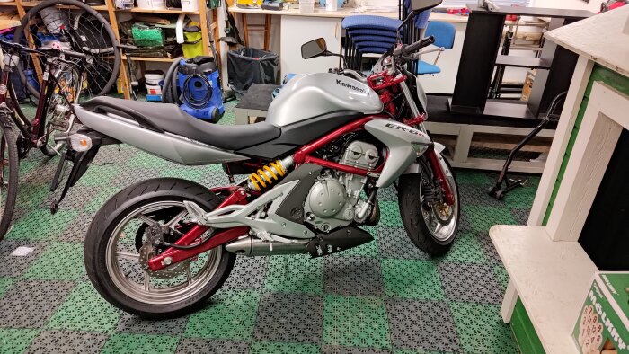 En silverfärgad Kawasaki ER-6n motorcykel står i ett garage, omgiven av cyklar och verktyg.