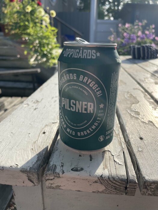 En burk Oppigårds Pilsner står på ett slitet träbord utomhus, med solbelysta blommor och en trädgård i bakgrunden.