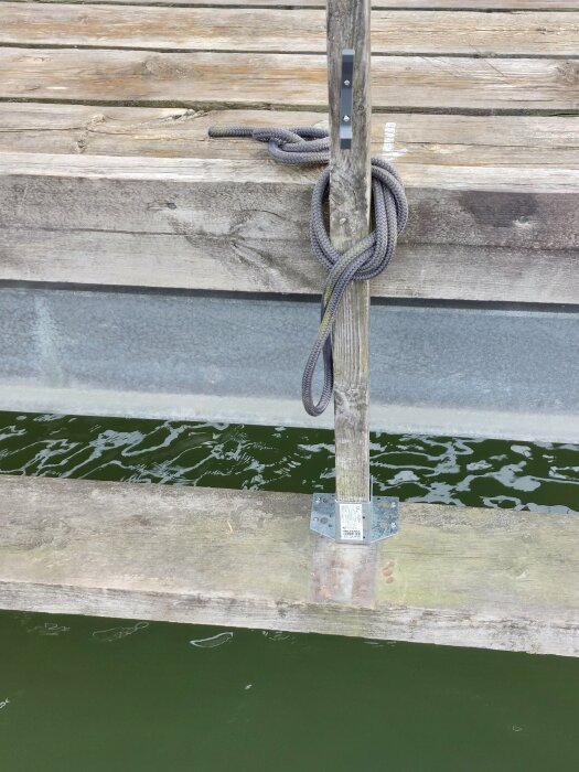 Trästolpe skruvad i bryggan, vid basen förstärkt med en balksko och fäst med träskruv, rep lindat runt stolpen; vatten och delar av bryggan synliga.