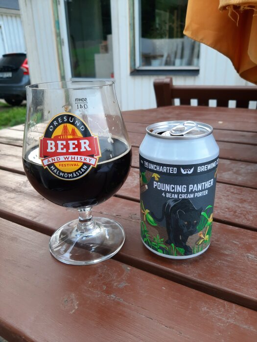 Ett glas mörk öl med etikett från Öresund Beer and Whisky Festival bredvid en burk Pouncing Panther 4 Bean Cream Porter sitter på ett träbord utomhus.