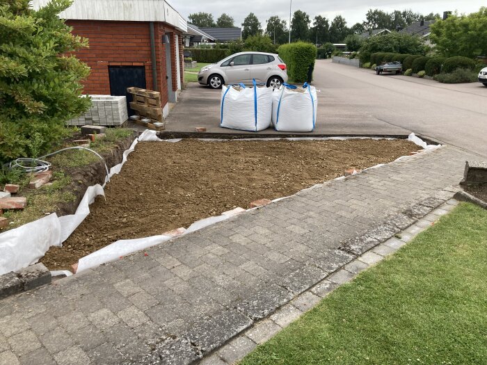 Uppgrävd yta framför ett hus förbereds med jord och makadam. Två stora säckar sand/grus på uppfarten. بریckor och pallar intill husväggen. Ytan omlindad med plastduk.