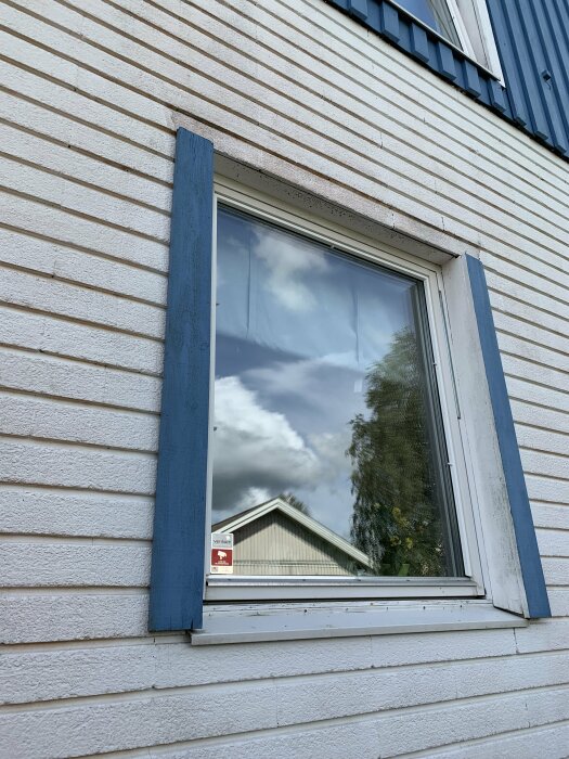 Närbild av ett fönster med blå fönsterfoder på en husvägg med vitmålad fasad. Skyltfönsterspegeln reflekterar himlen och ett grannhus.