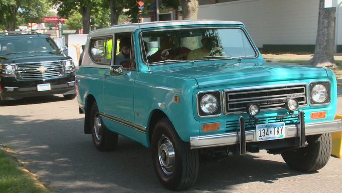 En ljusblå gammal SUV från 1979 körs fram på en gata, med en svart SUV bakom den. Träd och byggnader syns i bakgrunden.