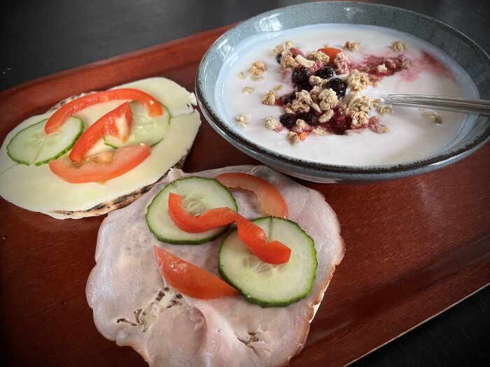 Frukostbricka med smörgåsar toppade med ost, paprika och gurka samt en skål yoghurt med musli och bär.