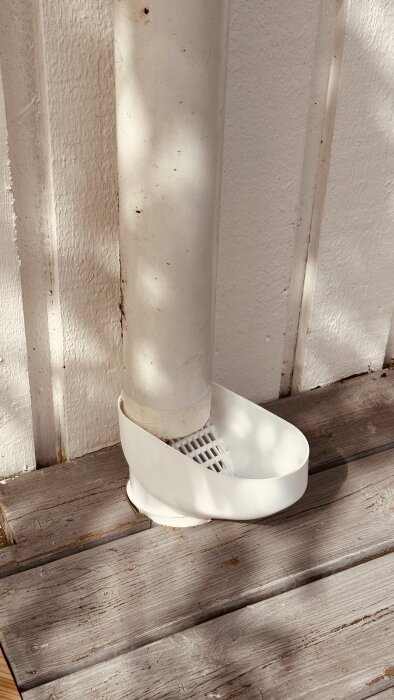 Ett vitt stuprör med en lövavskiljare monterad på terrassen bredvid en vit husvägg. Trallen har synliga borrade hål.