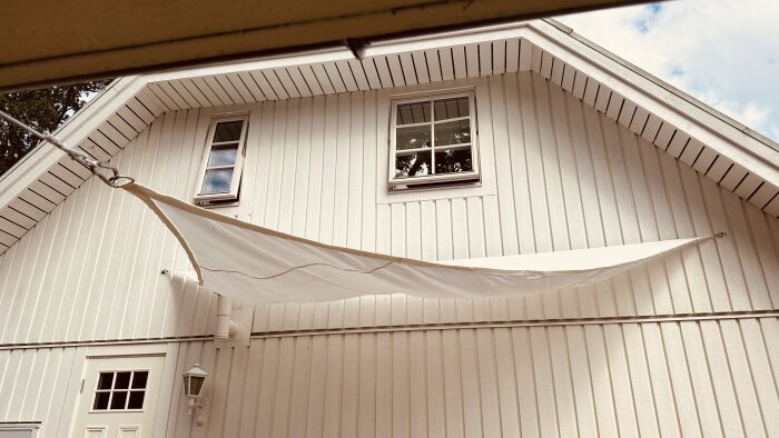 Vit trekantssegel spänd över en ny terrass, fäst i fasaden på ett vitt trähus med två våningar och små fönster.