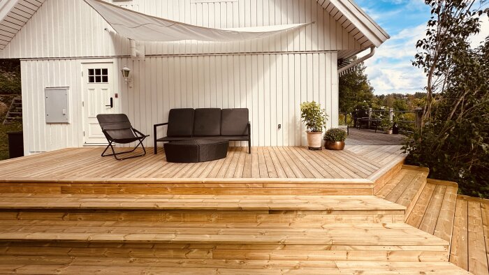 Nybyggd uteplats med trall, svart soffa och stol, runt bord, vita huset i bakgrunden, trekantssegel och två krukväxter.