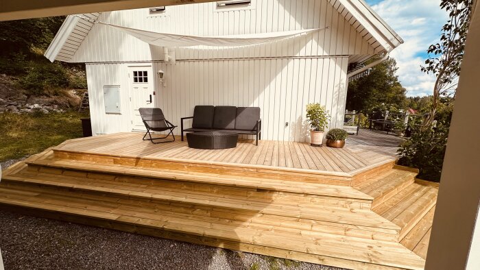 Nybyggd terrass i trä med trappsteg, en soffa, en plantkruka, en solstol och ett solsegel monterat ovanför, mot en vit träfasad.