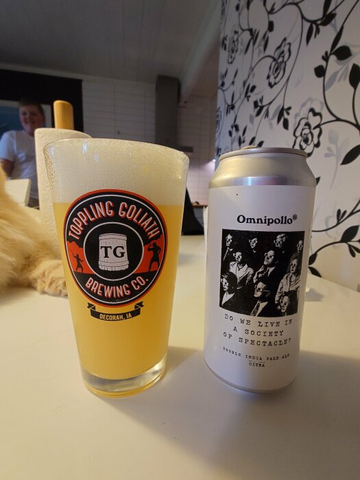 Ett glas öl från Toppling Goliath Brewing Co. och en burk Omnipollo Double IPA står på ett bord, med suddig bakgrund och blomtapet.