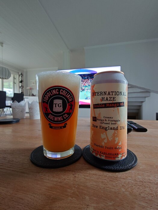 Ett glas och en burk öl av märket "International Haze" Double Mango Ed. New England IPA står på ett träbord i ett hem med en TV i bakgrunden.