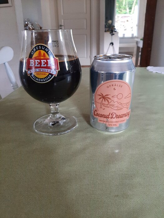 Ett glas med en mörk dricka från Öresund Beer Festival och en burk märkt "Ofrälse Coconut Dreaming Imperial Coconut Stout" på ett grön bord i en inomhusmiljö.