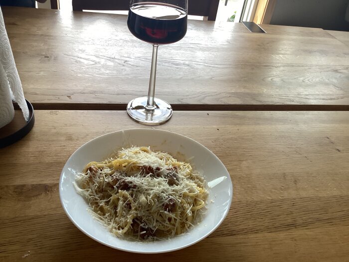 Tallrik med autentisk carbonara toppad med riven ost, bredvid ett glas rödvin på ett träbord.