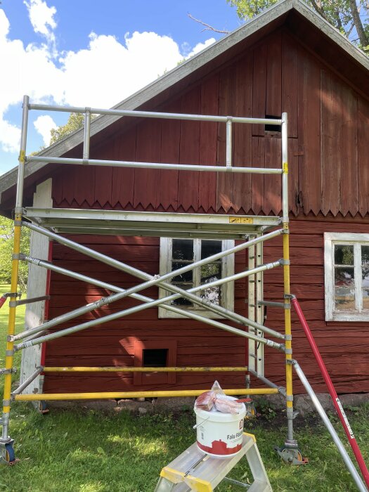 En röd byggnad med en ställning framför och en hink med färg märkt "Falu Rödfärg" på en pall, redo för målning. Klarblå himmel med vita moln i bakgrunden.