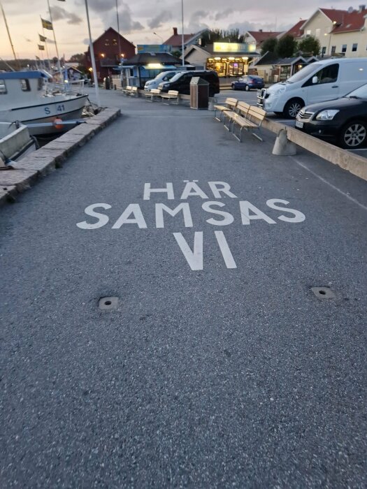 Gångväg vid marina med parkerade bilar längs kanten och texten "HÄR SAMSAS VI" målad på marken. Båtar och byggnader i bakgrunden.