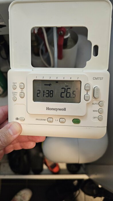 Hand som håller en Honeywell styrdosa/rumstermostat modell CM737, med en upplyst display som visar tiden 21:38 och temperaturen 26,5 grader samt en skiftnyckelikon.