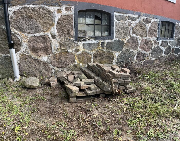 Stapel med stenar och tegelstenar vid sidan av en stenvägg med två små fönster; viss vegetation och ogräs på marken.