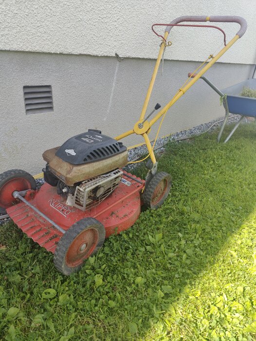 En gammal Klippo gräsklippare med en "Quattro 40" motor, ståendes på en gräsmatta bredvid en husvägg och en skottkärra i bakgrunden.